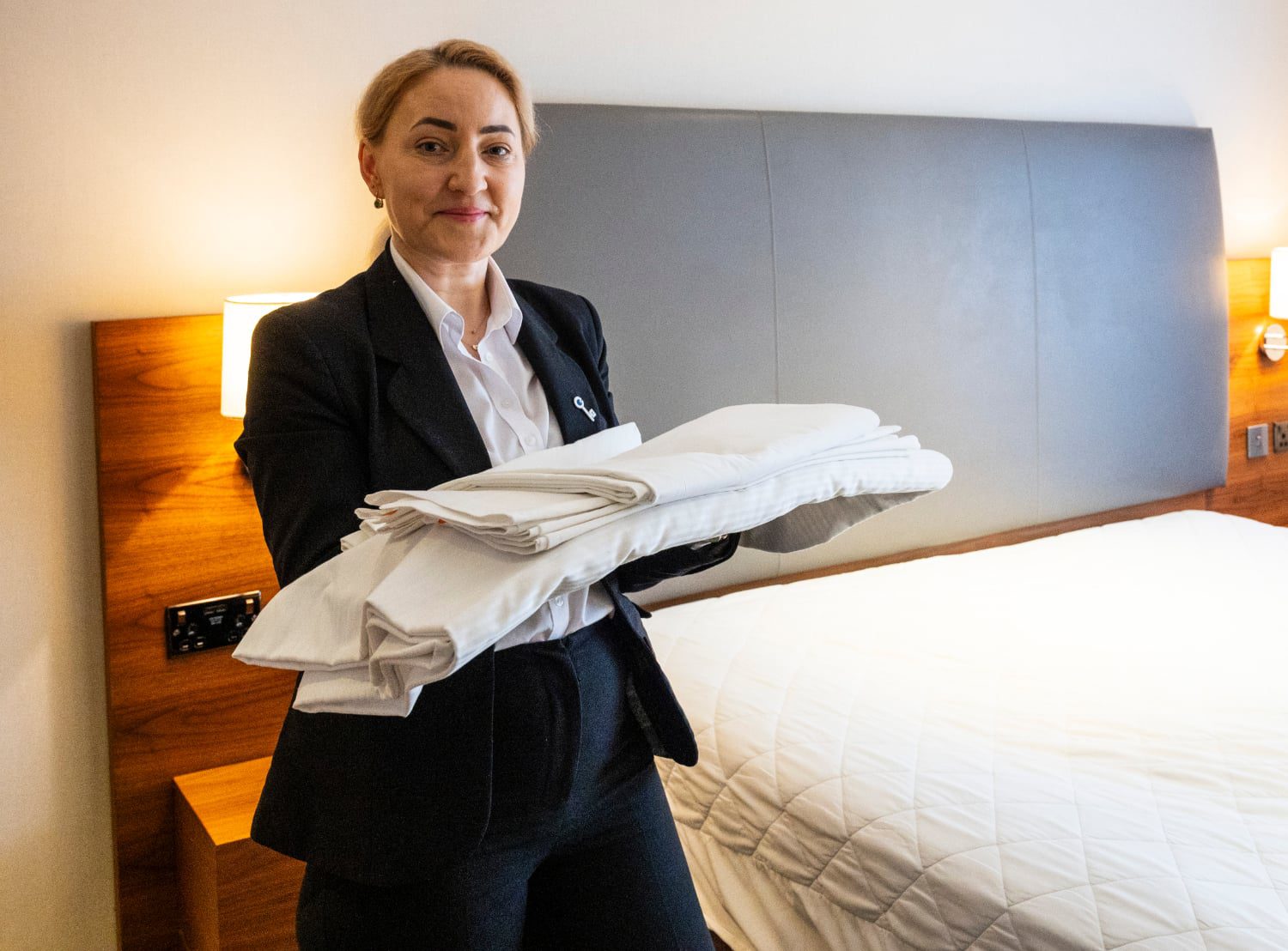 Hotel staff holding clean bed sheets in prepared room.